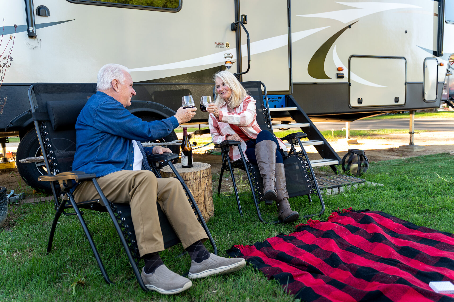 A senior couple seated clinking drinks, smiling and seated next to an RV Trailer.